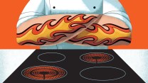 A chef stands in front of an electric stove with his arms folded over his chest. He has flame tattoos on this forearms, just over the electric stove.