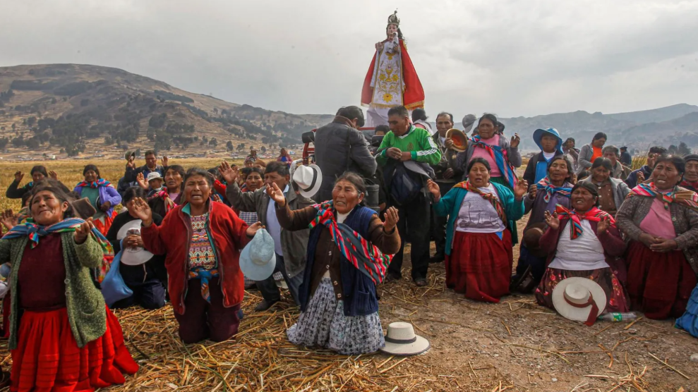 Community members kneel to pray on dirt and straw strewn ground. Mountains speckled with shrubs lay behind them.