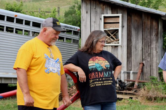 Jim, wearing a yellow shit, and Melissa, wearing a black shirt, standing outside some sheds. They are both white and are looking away from the camera.