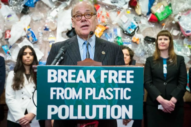 A white man wearing glasses and a blue tie stands at a podium with his mouth open. Behind him, several other individuals stand with serious faces. On the wall, there is a display of plastic trash.