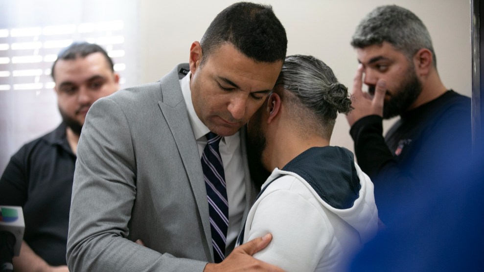 Two men, one in a grey suit, and one in a white hoodie, hug each other.