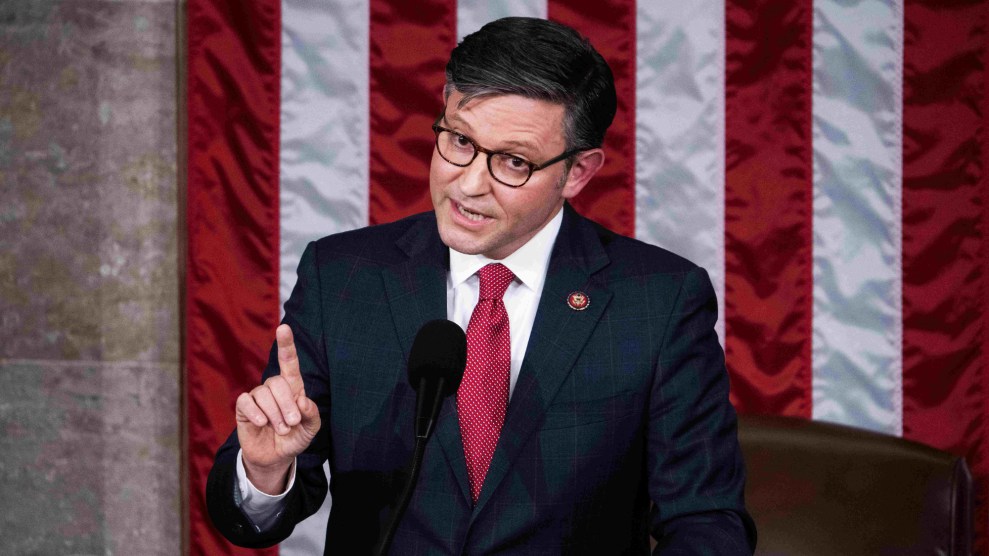 Mike Johnson is wearing glasses, a dark suit with a checkered pattern, a white shirt, and a bright red tie. He stands in front of the American flag, in front of a microphone, with one finger raised as if making a point or emphasizing something during his speech. He looks earnestly towards his audience, his fellow House members, from the elevated rostrum of the House of Representatives.