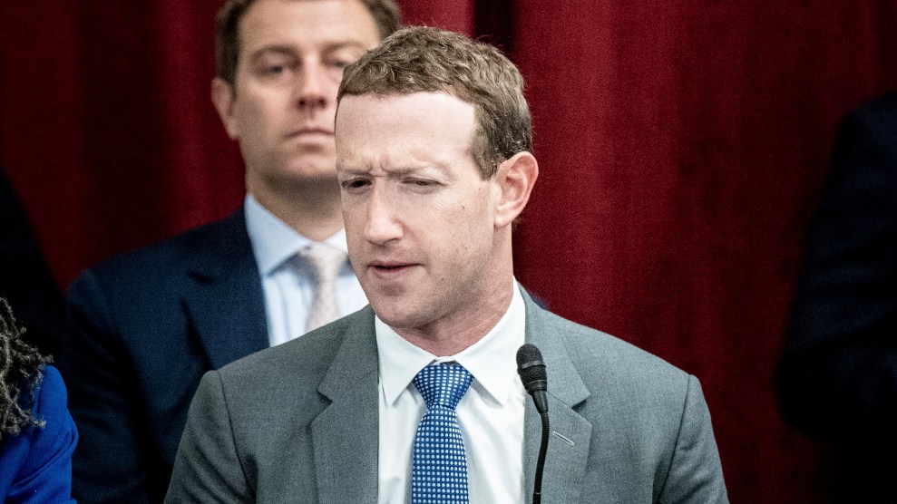 Mark Zuckberg sitting in front of a microphone, with his eyes squinting, in a suit, with red curtains behind him, and a man also seating directly behind him