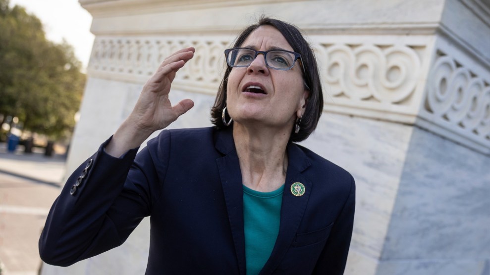 A brown haired woman stands in front of a marble monument with her her hand up and mouth open.