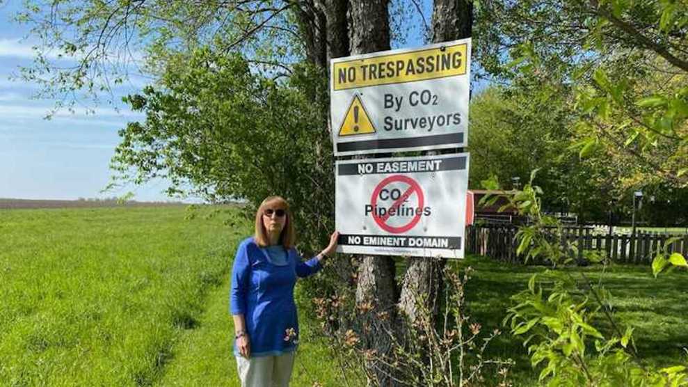 Kathleen Campbell, a white woman with red hair, standing next to signs on a tree that say "No Trespassing by CO2 Surveyors" and a red circle crossing out text that says "CO2 pipelines"