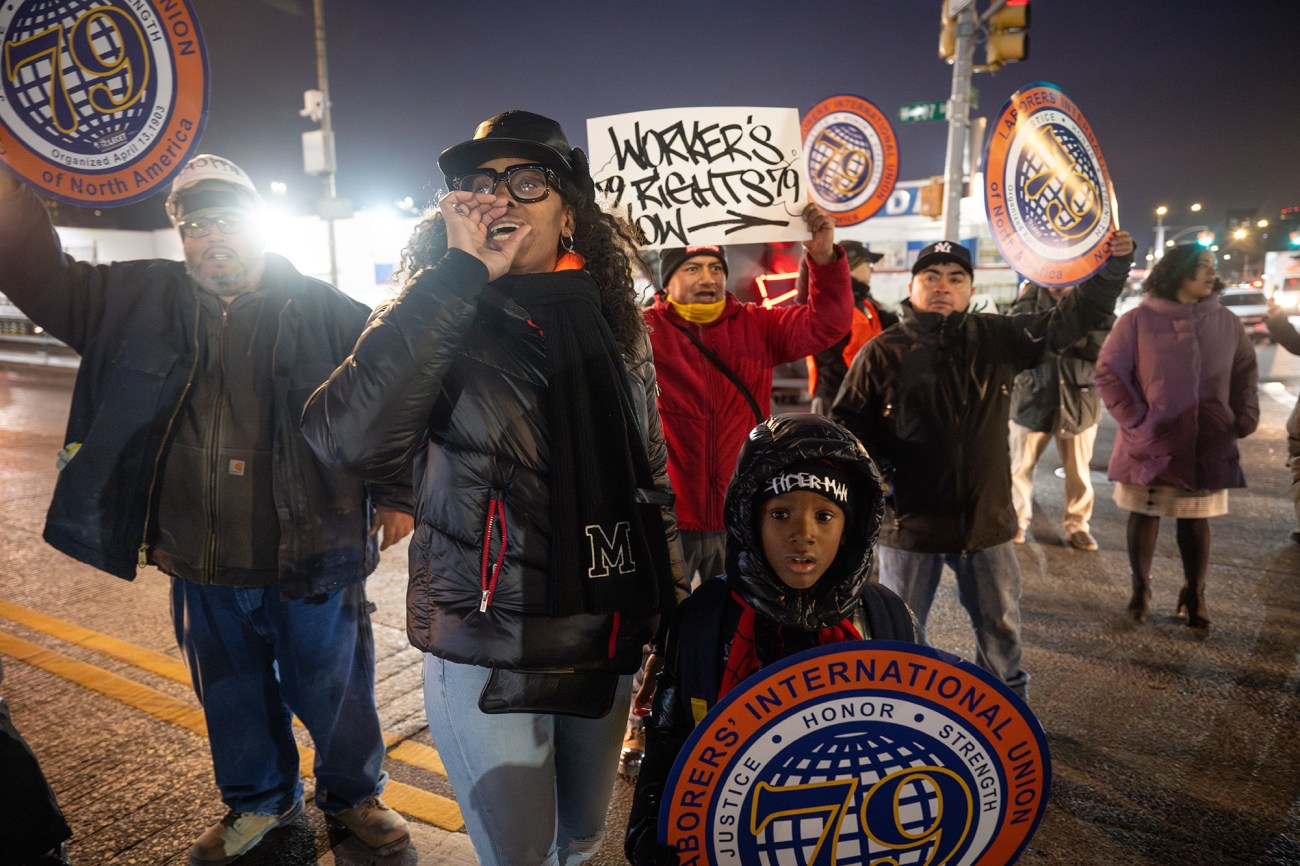 A woman stands in front of a group of people with protest signs, yelling.