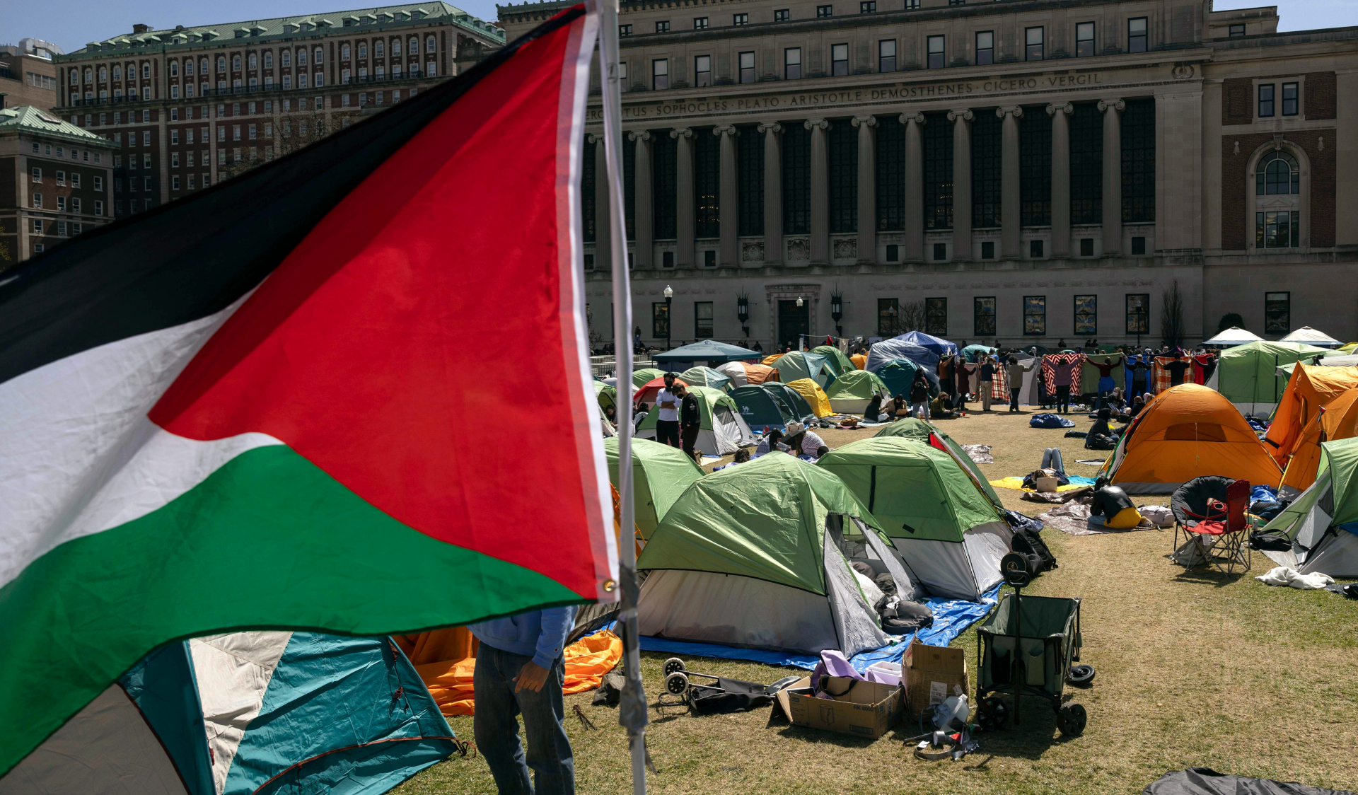 Tents fill a lawn outside a university building. A Palestinian flag flies in the foreground.