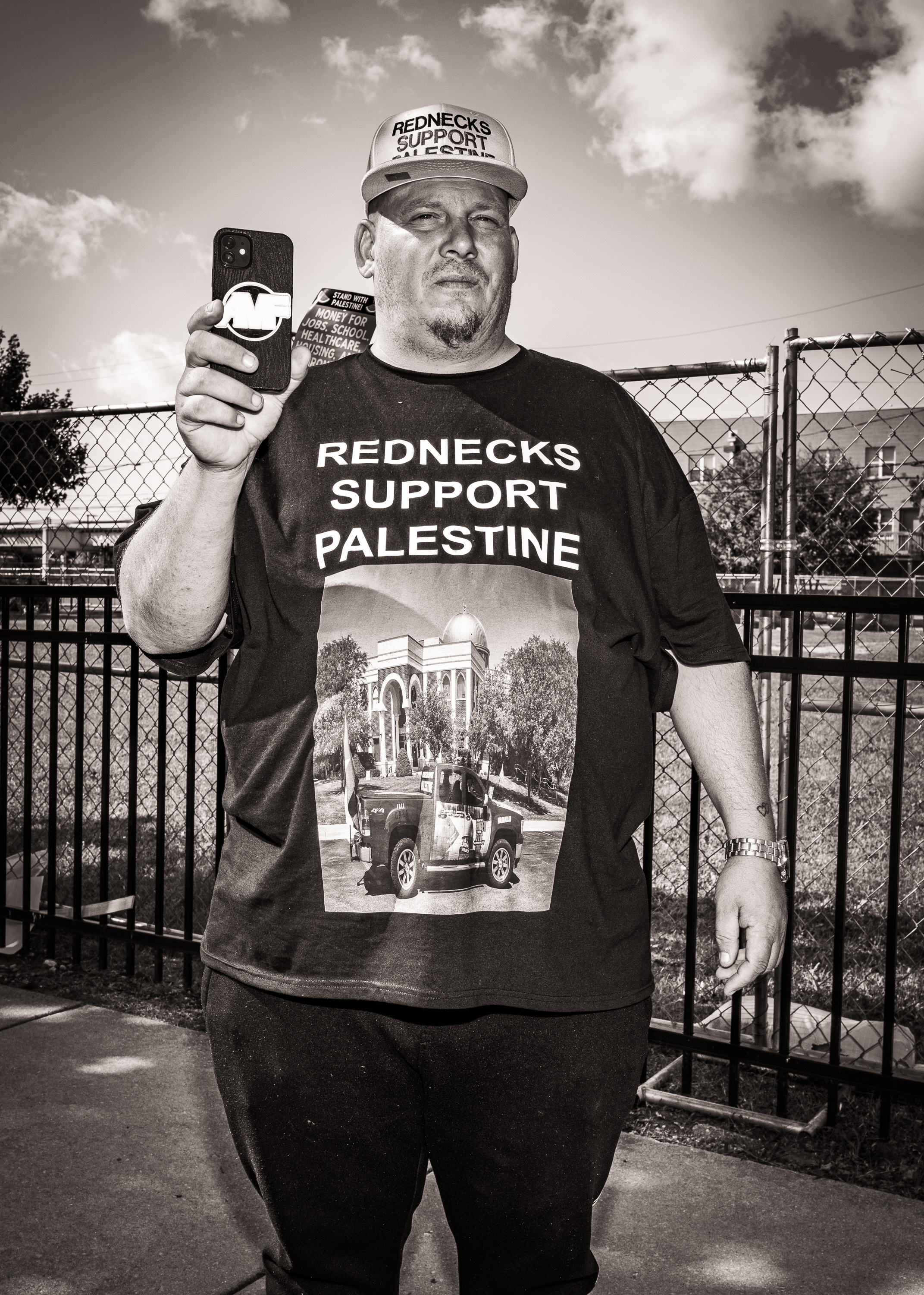 Black and white vertical photo of a man wearing a "Rednecks Support Palestine" t-shirt and hat, holding a cellphone.