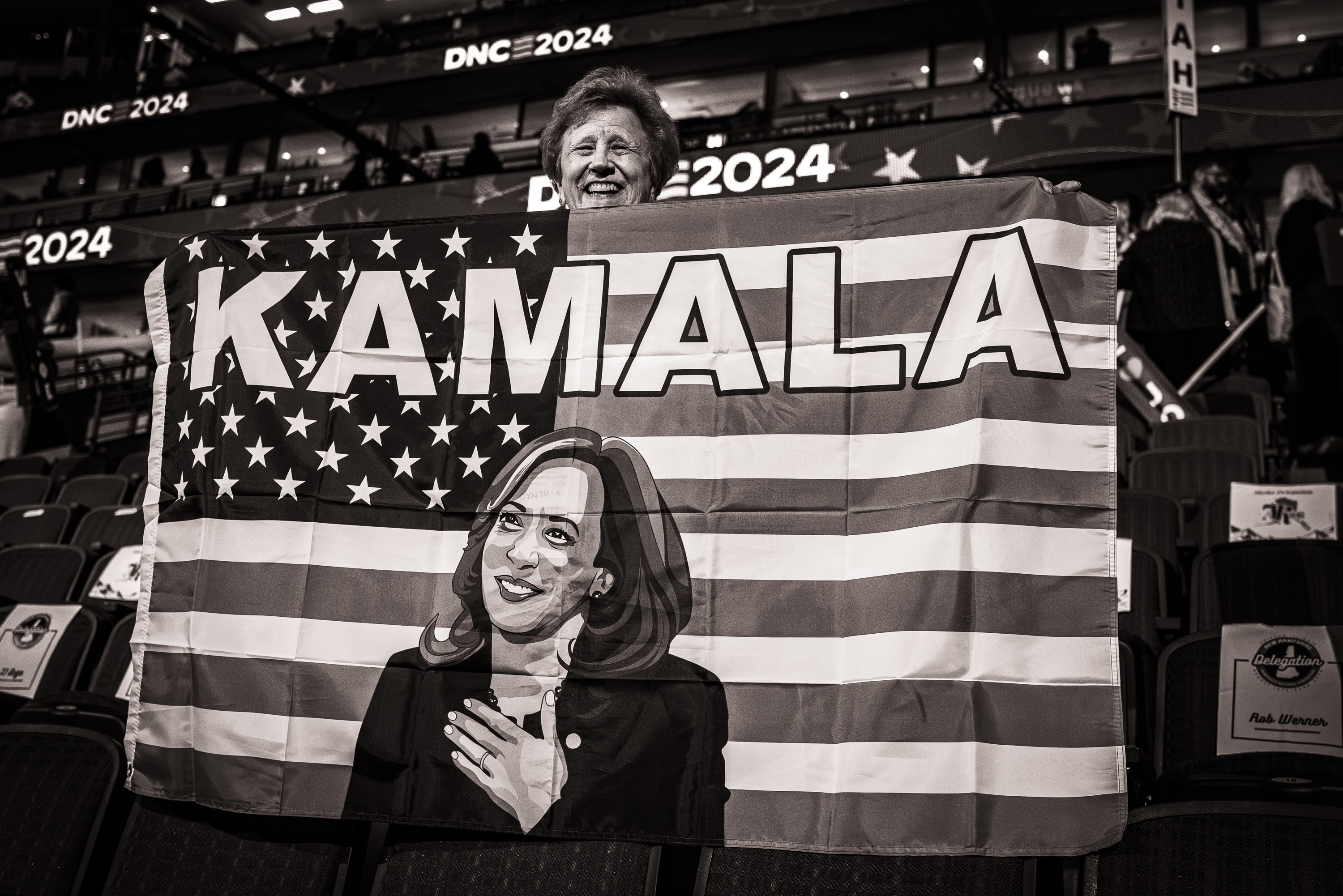 Black and white photo of a smiling woman holding an American flag that says "Kamala" at the top with an image of Vice President Kamala Harris on it.