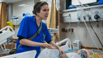 Dr. Tanya Haj-Hassan, in blue scrubs, holds the fingers of an injured child lying in a hospital bed.