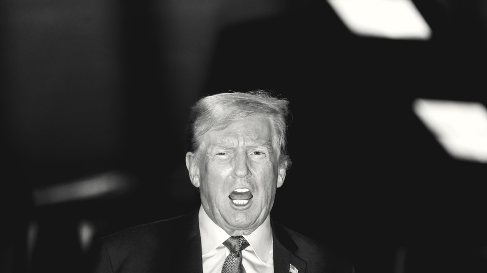 A black and white photo of Donald Trump speaking to the media. The photo is taken using a noticeable flash, which casts Trump's shadow against the ceiling.