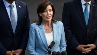 New York Gov. Kathy Hochul, a white woman with brown hair, wearing a light blue suit. She is speaking into a microphone and has two men, one white and one black, behind her in suits.