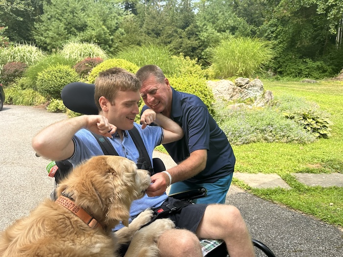 A white aging man kneeling near his son, a younger white man in a wheelchair, with a golden retriever also leaning towards a man in a wheelchair