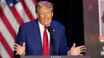 Donald Trump speaking into a microphone in a white shirt, blue shirt and red tie. His hands are outstretched to the side and he has a notable spraytan