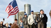 Two men wearing blue "America First" hats with a US flag in front of General Motors' headquarters.