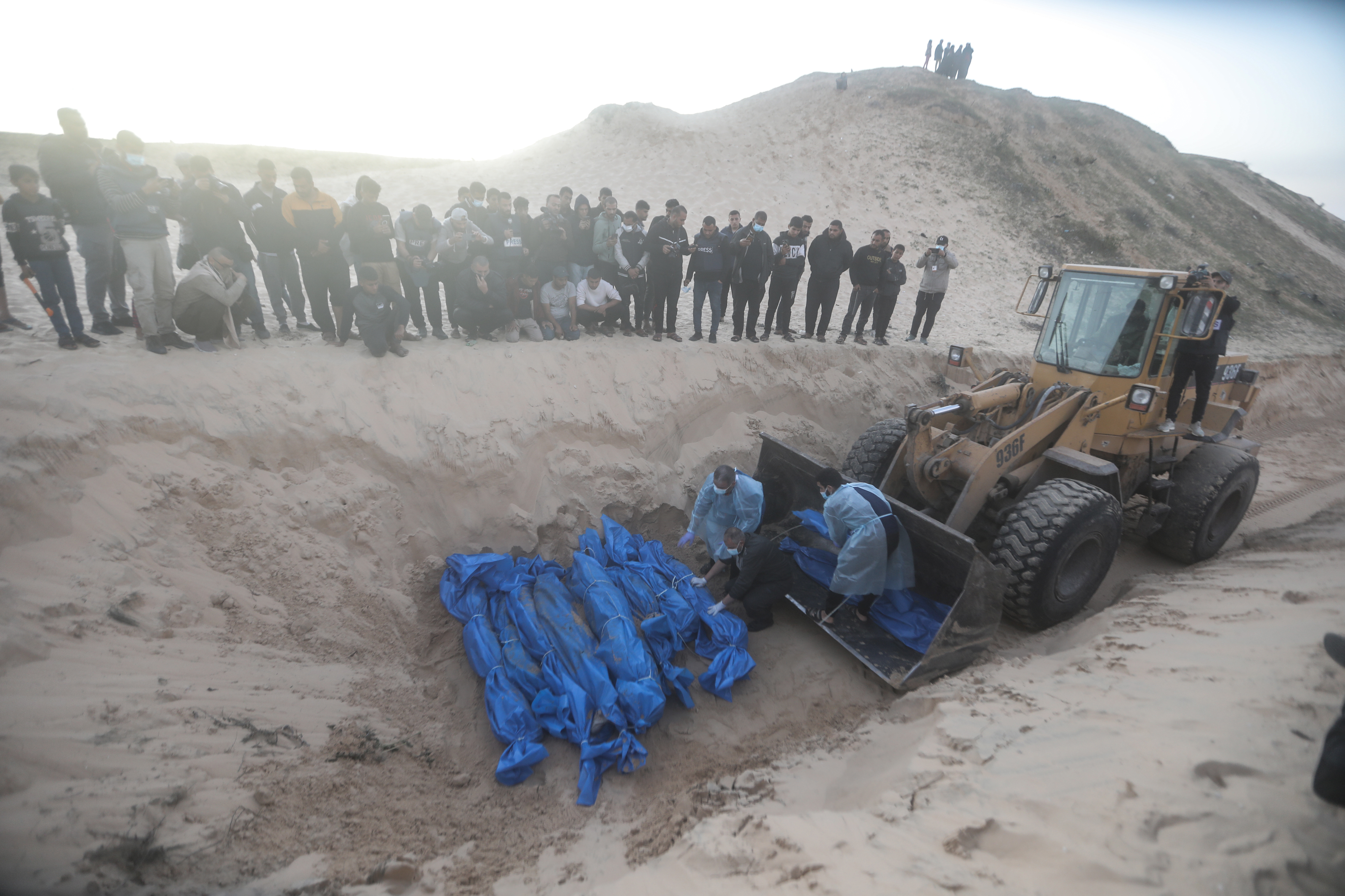 A bulldozer pushes dirt over a mass grave as mourners look on. 