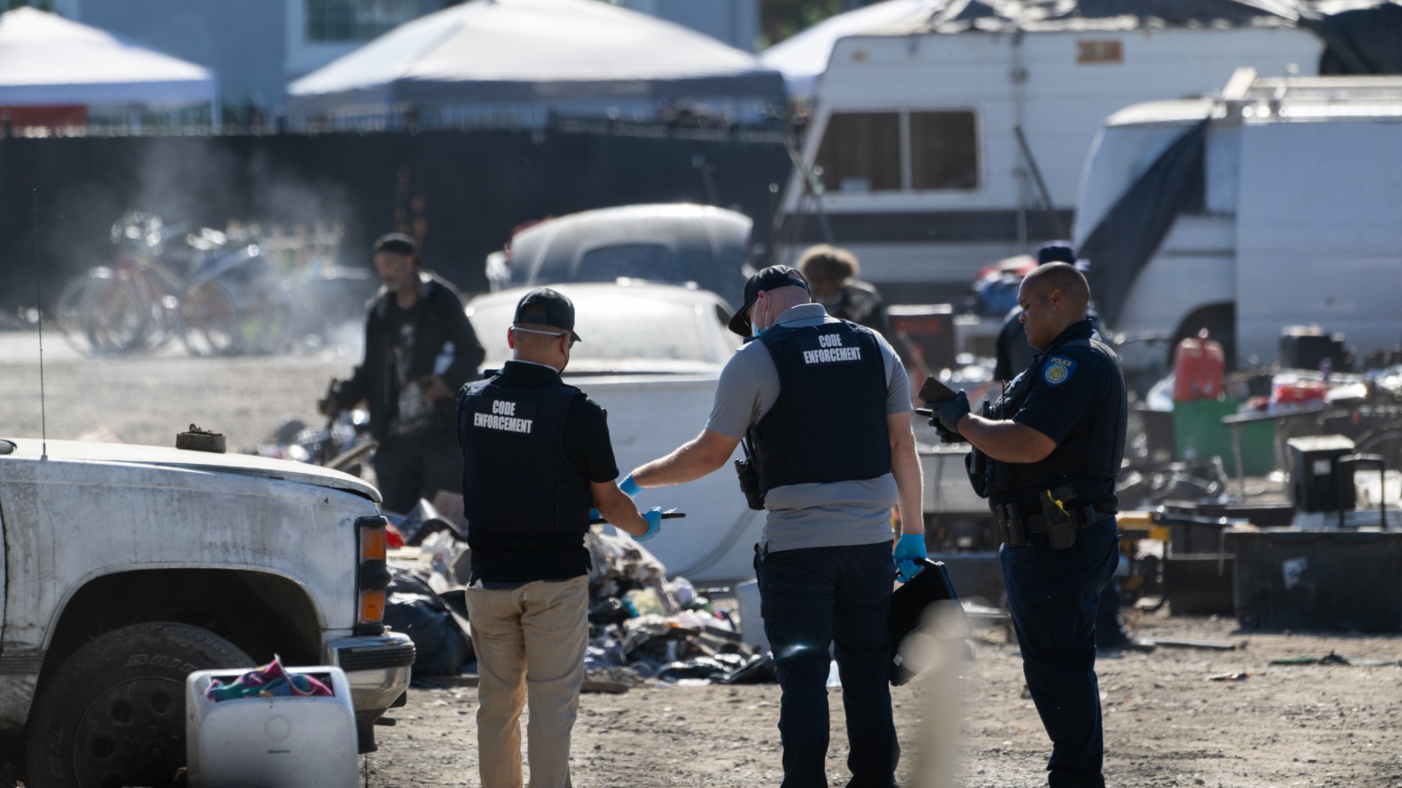 Two code enforcement officials and a police officer oversee the clearing of a homeless encampment.