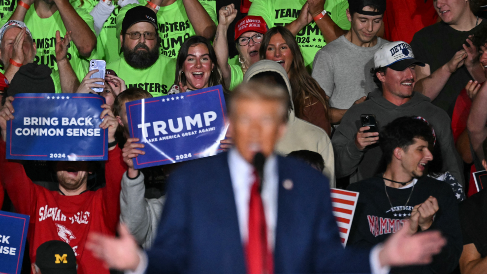 A photo of out-of-focus Donald Trump standing behind the podium, speaking to the crowd at a rally. Behind him, in focus, are people with pro-Trump signs, wildly cheering.