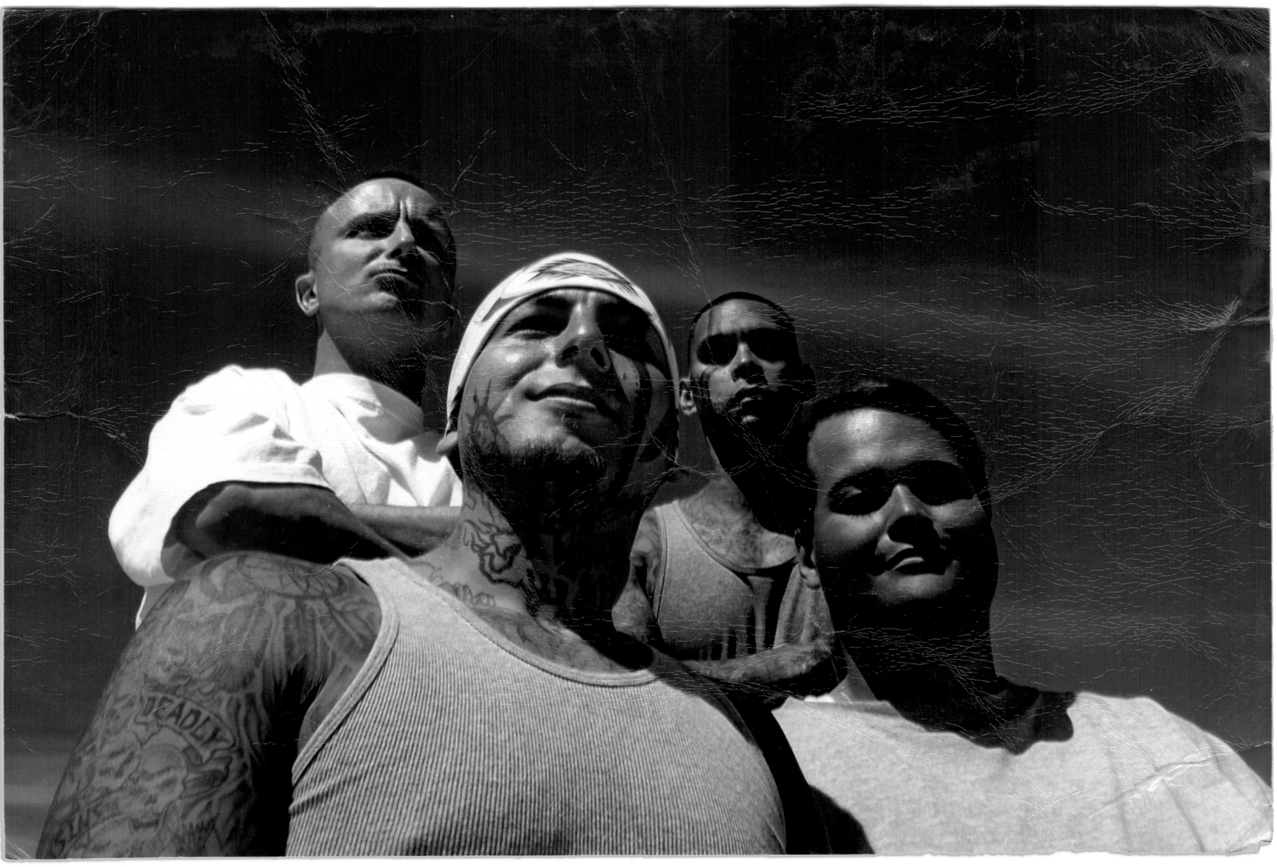 In a black and white photograph, Grace Pinson, a trans Latina with short dark hair, stands outside in the sun with three men. The person to her left wears a tank top and has tattoos on his face, neck and arms. Behind her stand two men. The one on the right wears a white T-shirt and is bald. The one on the left has tattoos on his chest and arms and has close-cropped, dark hair.