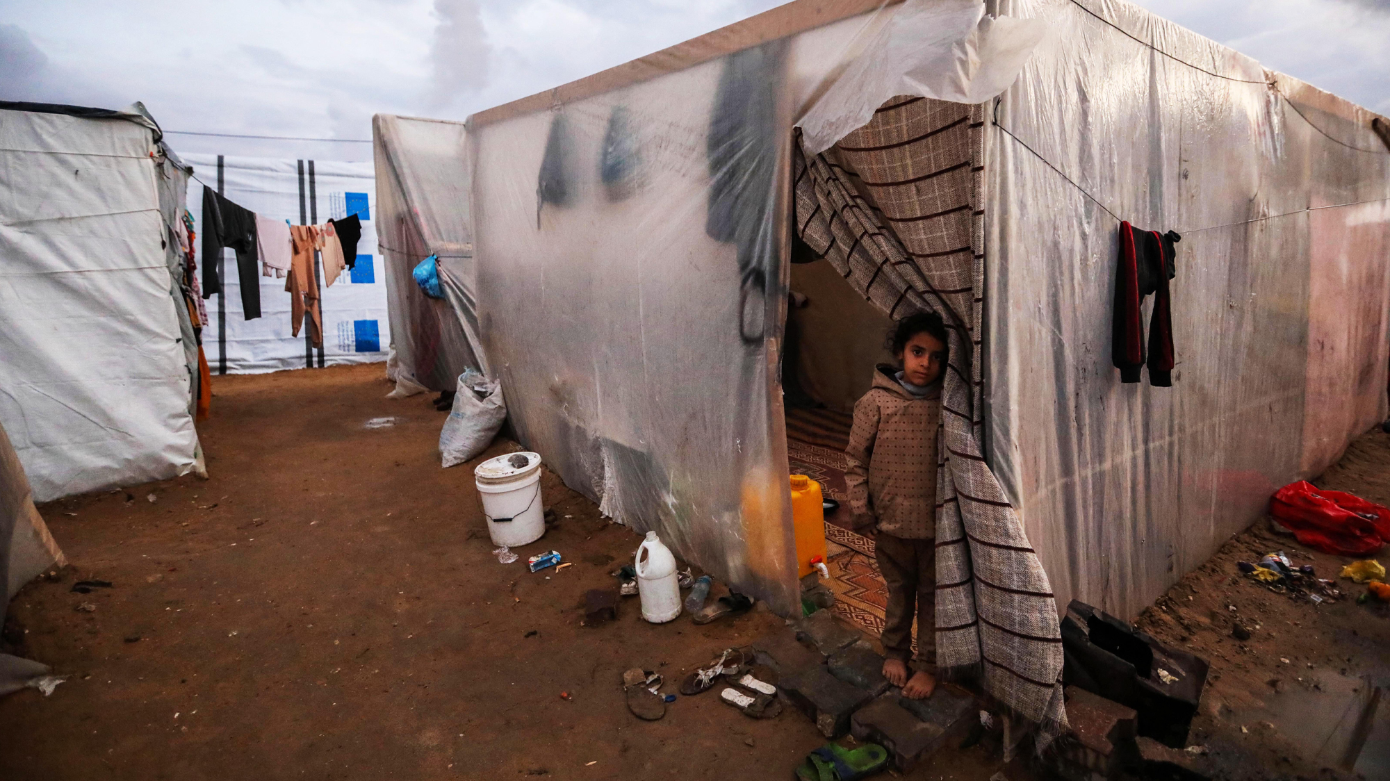 A barefoot Palestinian girl stands in the doorway of a plastic-walled living space in a squalid temporary camp, with trash, plastic jugs, and buckets on the ground and clothes hanging from clotheslines.