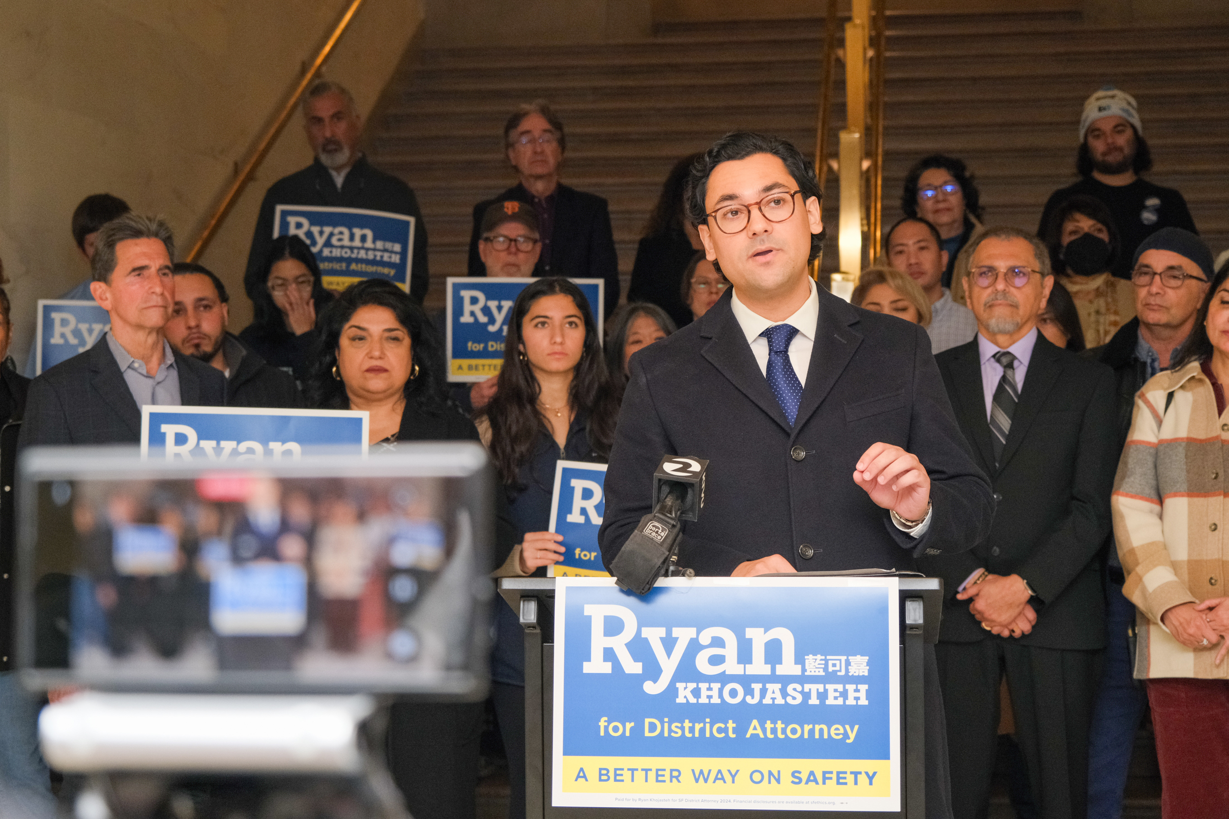 Ryan Khojasteh, wearing glasses along with suit and tie, gives a speech behind a podium. On the front of the podium is a campaign sign that reads: Ryan Khojasteh for District Attorney: A Better Way on Safety. About 20 people stand behind him in support of his candidacy.