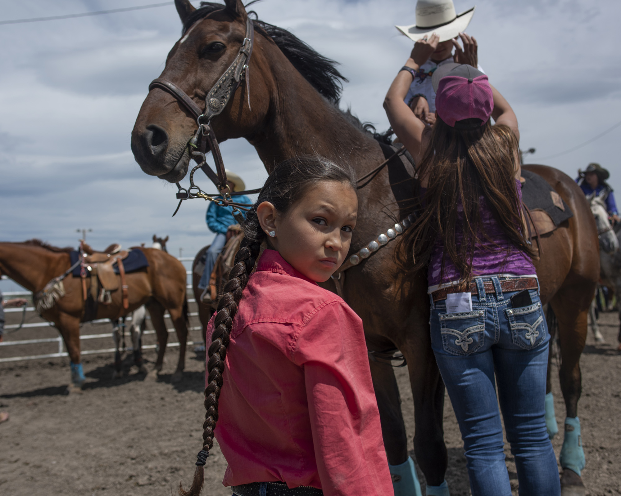 A girl with a long braid looks at the camera, standing in front of a saddled horse with a rider.