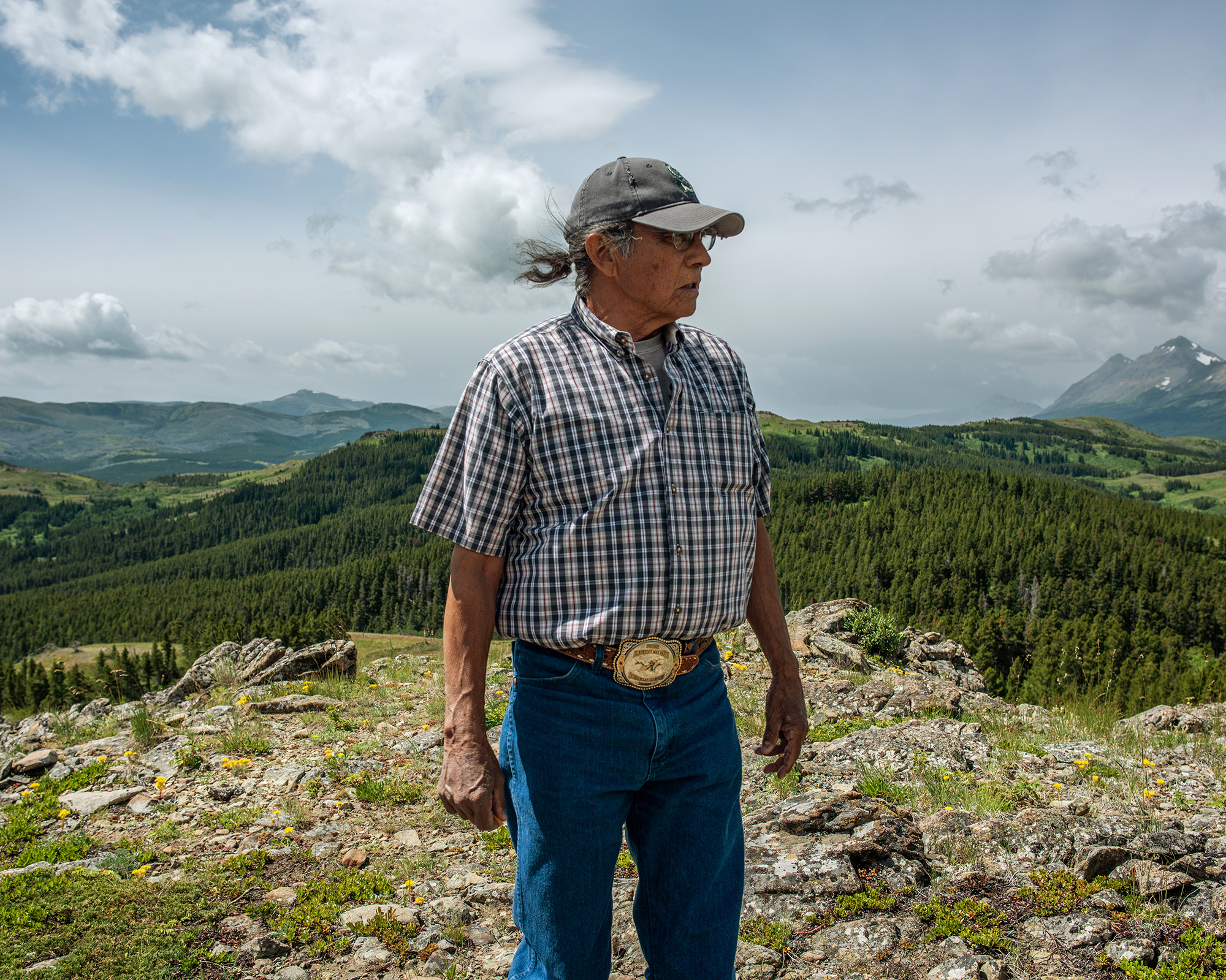 Portrait of a man wearing a baseball hat and large belt buckle, standing on a mountaintop area.