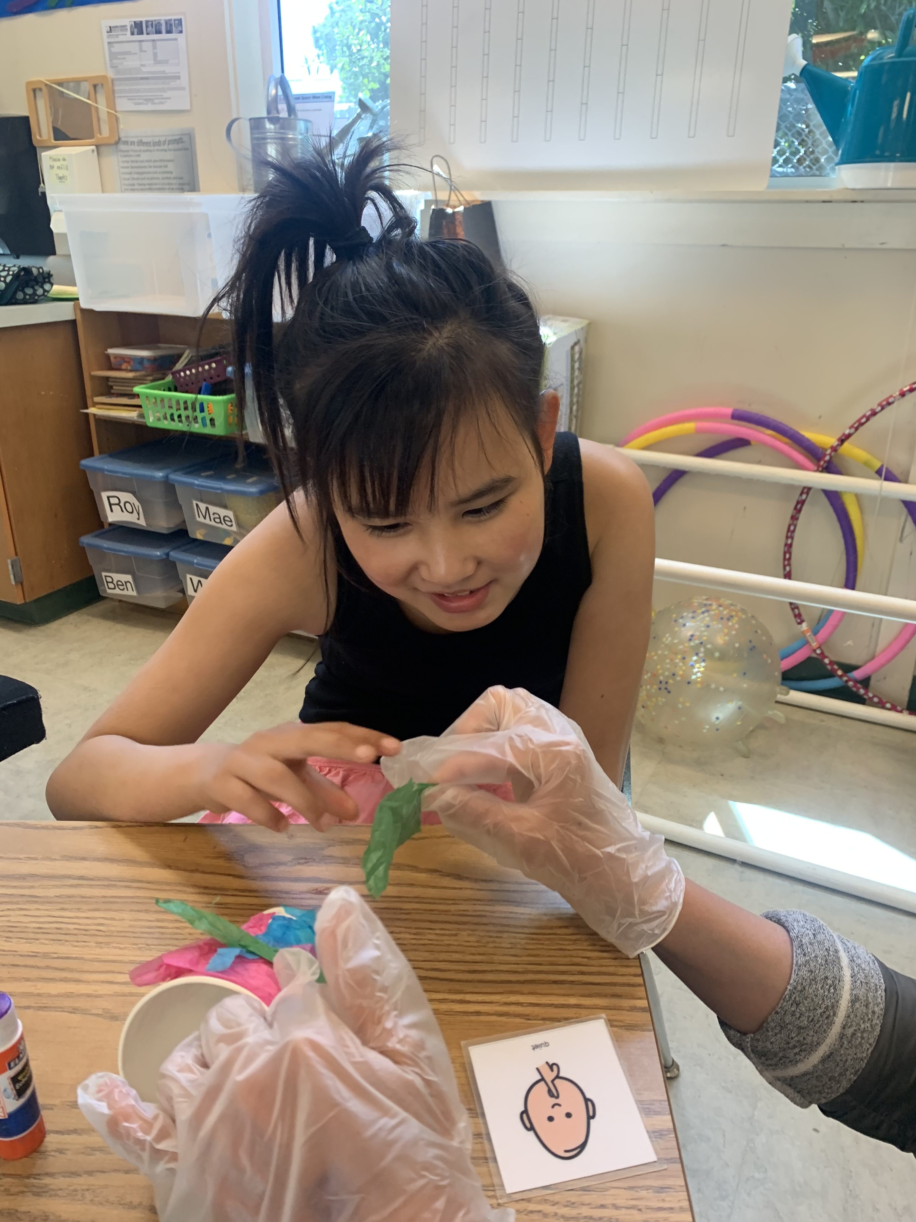 A girl works on an art project involving tissue paper with an aide, who wears plastic gloves.