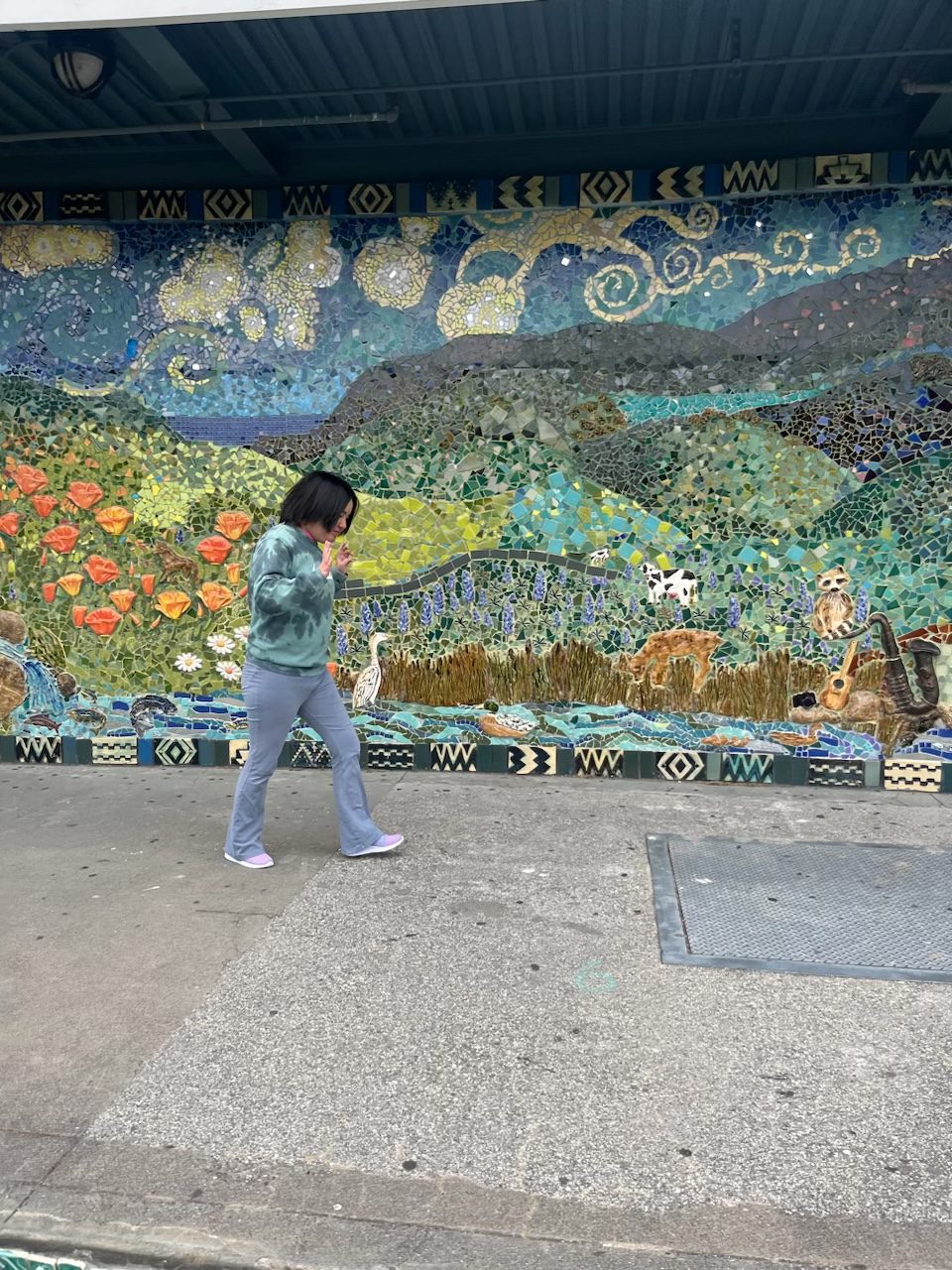 A teen girl walks down a sidewalk with a large tile mosaic of a nature scene behind her.