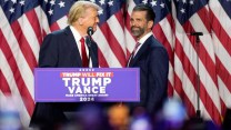 Donald Trump in a dark suit and red tie stands behind a campaign podium with his son Don Jr. against several large American flags in the background