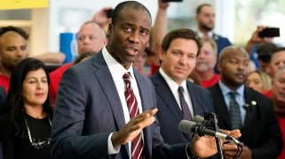 Joseph Ladapo stands at a microphone. Ron DeSantis and others in a crowd stand behind him.
