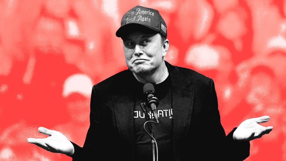 Elon Musk, dressed in all black with a black 'Make America Great Again' hat shrugs on stage in front of a microphone.