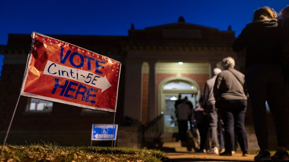 A photo of a dozen voters stand on a walkway outside the Cincinnati Observatory, which is a Greek Revival style building. The people are lit up by a street lamp as they've arrived in the early morning hours. In the grass beside the walkway are two signs. The red sign with blue type reads: Vote Here: Cinti 5E and it has a white arrow pointing toward the door. A blue sign in the distance has a white arrow that points the opposite way and reads "Handicapped Access."