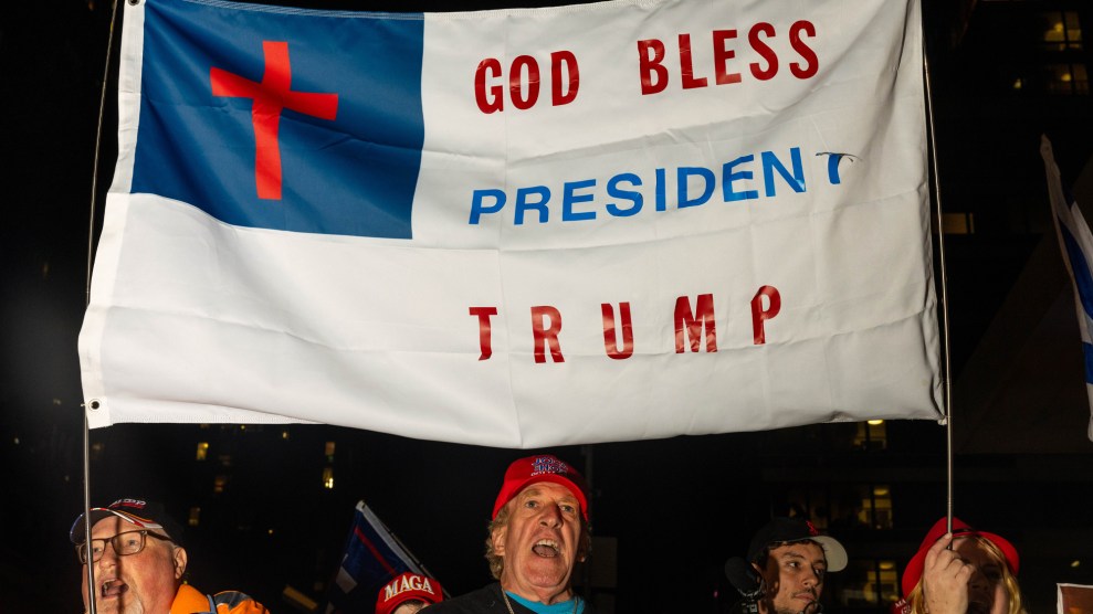 A photo of several men marching down the street at night, chanting. The men are wearing red hats that read MAGA and the men are holding poles that raise a Christian flag that reads "God Bless President Trump."