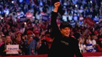 Elon Musk holds his fist in the air at a campaign rally for then Republican presidential nominee Donald Trump at Madison Square Garden in October. Musk, dressed in a black cap, black blazer and black t-shirt, smiles in front of a crowd of people in red and blue taking photos and holding signs behind him.