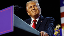 Donald Trump in blue suit and red tie, leaning on a lectern. He has a big closed-mouth smile on his face, as he looks to the right.