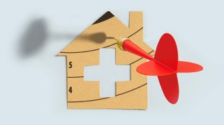 An illustration of a cardboard cutout shape of a house. The shape of the house has been cut from a dartboard pattern, and at the center of the house is the international aid sign. A red dart with a gold tip pierces the roof of the house.