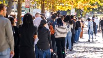 A group of people standing in a long line outside, with them facing away from the camera