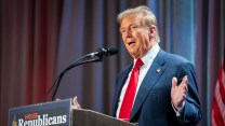 Donald Trump stands on stage behind a podium, arms spread out, mid sentence