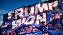 A person in a US flag sweatshirt stands in front of a massive flag that says, "Trump Won."