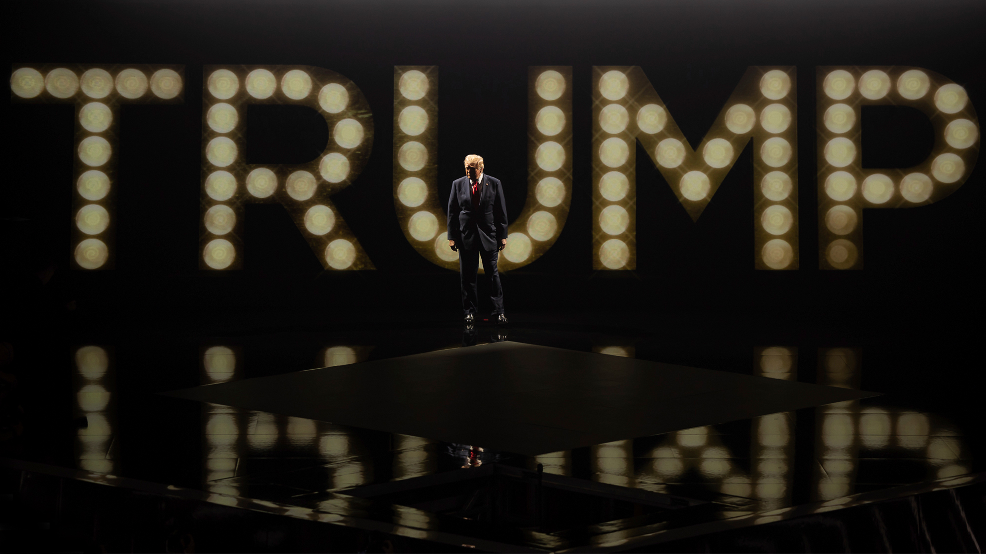 Donald Trump stands on a darkened stage, backlit by his name "TRUMP" spelled out in large lights.