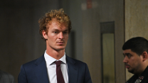 A young caucasian man with curly hair, wearing a suit and tie, stares blankly as he walks past a police officer in the hallways of the court building.