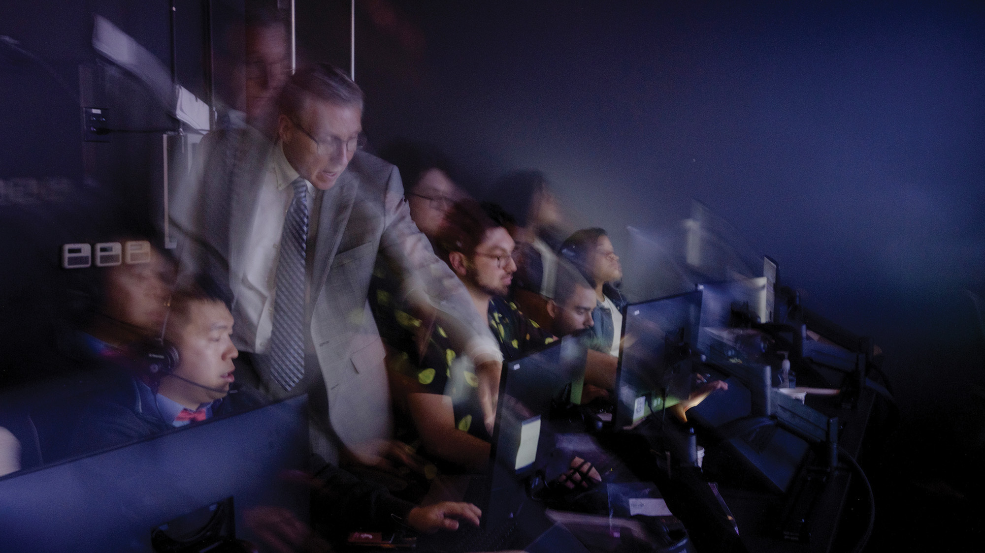 Motion-blurred image of several people sitting in front of computer terminals in a dark room.