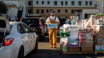 A bearded young man wearing light brown overalls and a blue Los Angeles Dodgers baseball cap carries a package of bottled water. To the left of the man are a line of cars. To the right of the man are palettes of packaged goods, such as pampers and bottled drinks.