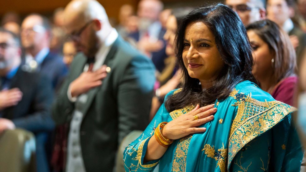 A woman places her right hand over her heart. She's wearing a teal salwar kameez embroidered in gold patterns.