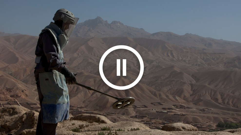 A pause symbol overlaid on a photograph of a deminer uses a metal detector to search for unexploded weapons in the mountains of Afghanistan.