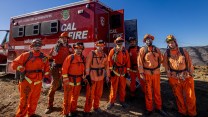 Seven people in bright orange jumpsuits stand in front of a fire truck.