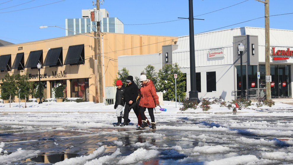 A group of three people bundled in jackets walk through an intersection covered in snow. The sky is blue behind them.