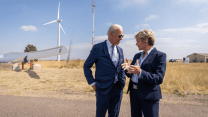 A white man and a white woman both wearing suits talk to each other in front of a windmill in a desert.