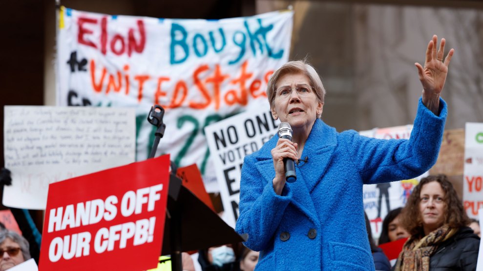 A crowd of people with signs and banners stand behind Elizabeth Warren, who has short-cut hair and wears thin-rimmed glasses. Dressed in a blue winter coat, she holds her left hand up to the crowd as she speaks into the microphone in her right hand. The red sign on the lectern in front of her has white letters that reads, "HANDS OFF OUR CFPB!"